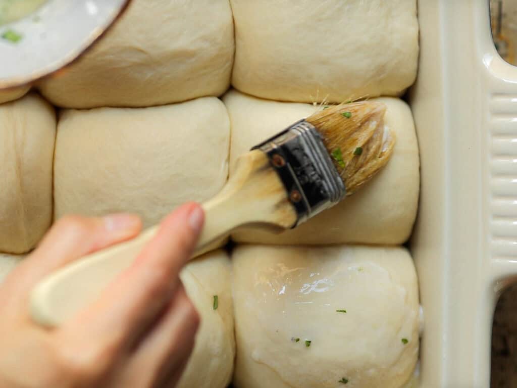 Perfect Roasted Garlic Vegan Dinner Rolls Recipe. 34 A hand brushes melted butter with herbs onto unbaked dough rolls in a baking dish, preparing them for baking. The rolls are neatly arranged in rows.