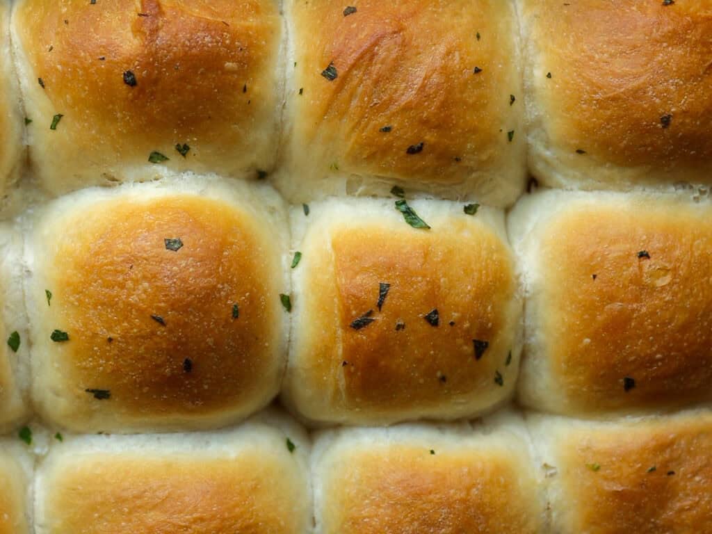 Perfect Roasted Garlic Vegan Dinner Rolls Recipe. 35 Close-up of freshly baked, golden-brown vegan dinner rolls topped with small flecks of herbs. The rolls are arranged in a grid pattern, showcasing their soft texture and fluffy appearance.