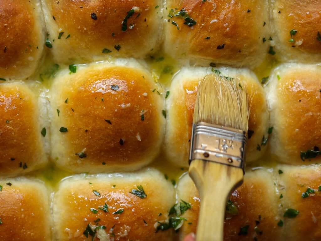Perfect Roasted Garlic Vegan Dinner Rolls Recipe. 36 A close-up of golden-brown vegan dinner rolls being brushed with melted garlic butter, garnished with chopped parsley.