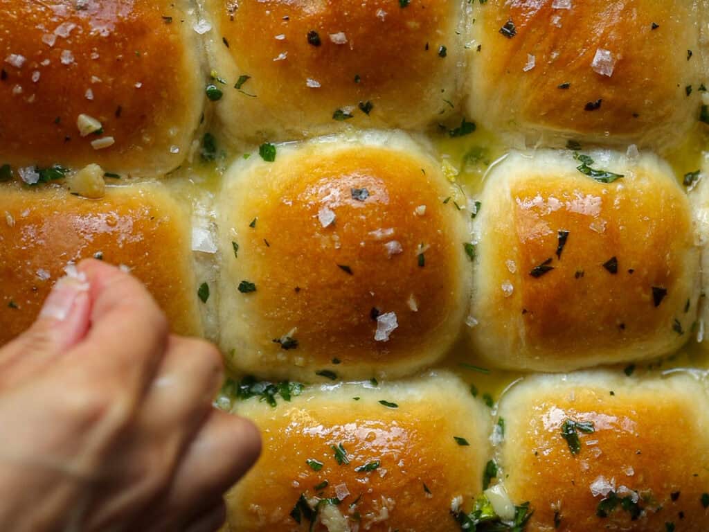 Perfect Roasted Garlic Vegan Dinner Rolls Recipe. 2 A hand sprinkles sea salt over freshly baked vegan dinner rolls glazed with melted butter and garnished with chopped herbs. The golden brown tops of the rolls are shiny and appetizing.