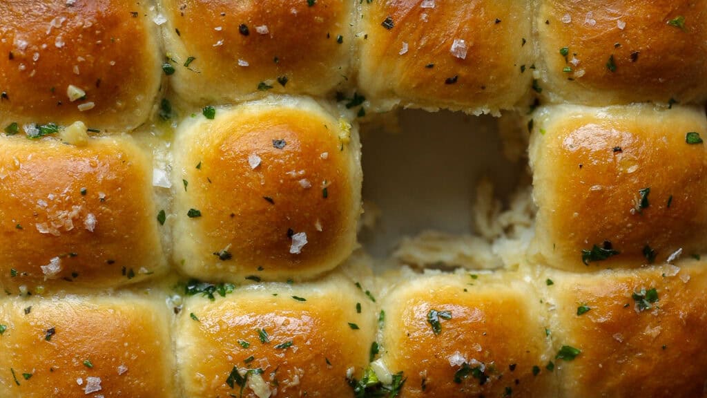Perfect Roasted Garlic Vegan Dinner Rolls Recipe. 1 Golden-brown garlic vegan dinner rolls arranged in a grid with one missing piece, topped with herbs and sea salt, showcasing a soft, fluffy interior from a birds-eye view.