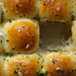 30+ AMAZING VEGAN Holiday Recipes Everyone's gonna Love. 4 Close-up of golden-brown garlic vegan dinner rolls arranged in a grid pattern. Center roll is missing, revealing soft, fluffy interior. Sprinkled with herbs and salt, the bread features a shiny surface characteristic of melted butter.