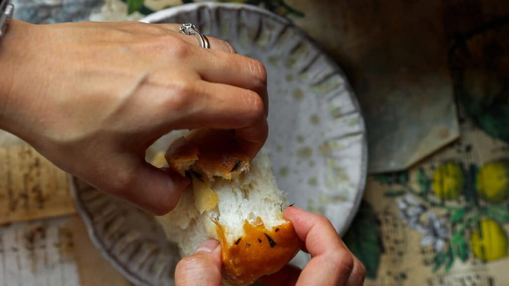 Perfect Roasted Garlic Vegan Dinner Rolls Recipe. 38 Two hands breaking a piece of bread above a decorative plate. The table underneath has a floral and lemon pattern. The person wears a ring on their right hand.