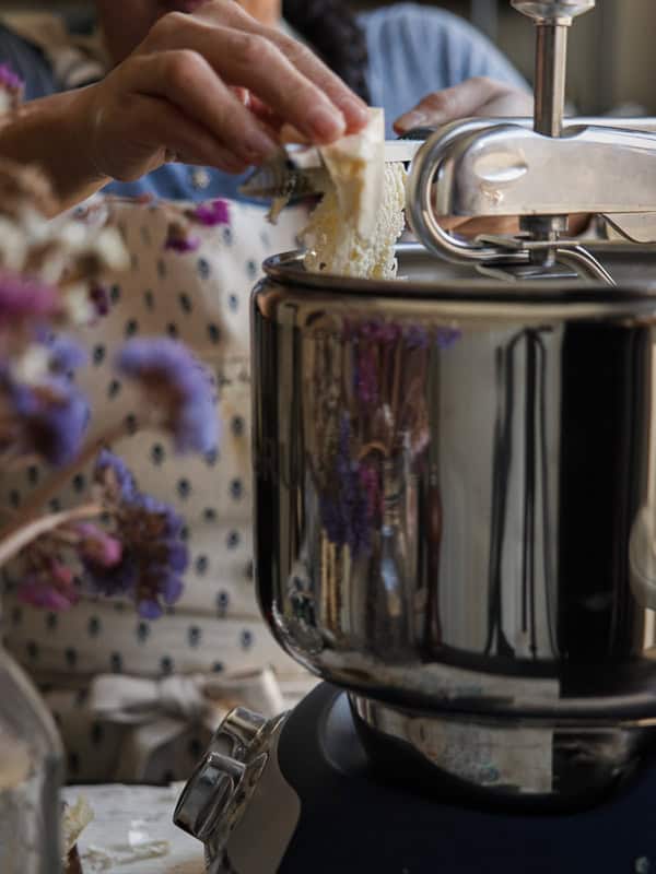 Perfect Roasted Garlic Vegan Dinner Rolls Recipe. 17 A person grates vegan butter into a stand mixer. They wear a dotted apron, and dried flowers are visible in the foreground.