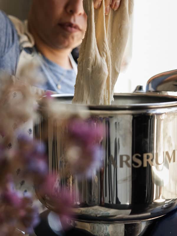 Perfect Roasted Garlic Vegan Dinner Rolls Recipe. 20 A person kneads dough with both hands in a shiny metal mixer. Flowers are blurred in the foreground, adding a touch of color to the scene. The focus is on the dough stretching out from the mixer.