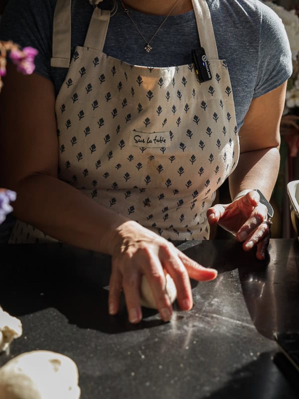 Perfect Roasted Garlic Vegan Dinner Rolls Recipe. 29 A person wearing a patterned apron is kneading dough on a dark countertop in a sunlit kitchen. Their hands are positioned over the dough, and they are wearing a smartwatch on their left wrist.