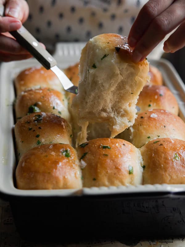 Perfect Roasted Garlic Vegan Dinner Rolls Recipe. 3 A hand is pulling a fluffy, golden brown dinner roll garnished with herbs from a baking dish using a knife. The rolls are shiny and appear freshly baked with a soft texture.
