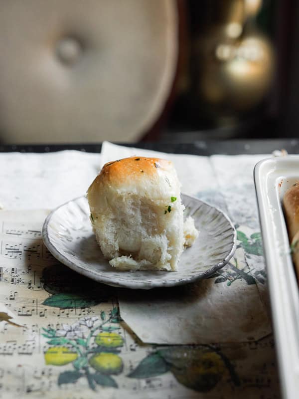 Perfect Roasted Garlic Vegan Dinner Rolls Recipe. 37 A single soft dinner roll with a golden top sits on a small, textured ceramic plate. The background includes a botanical illustration and a hint of musical notes on paper. The setting is cozy, with muted lighting.