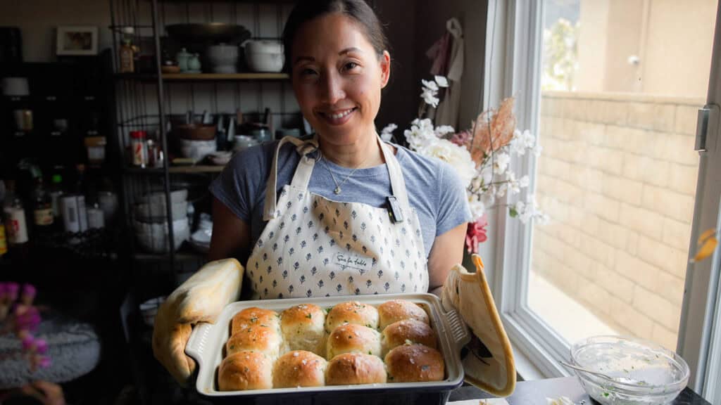Perfect Roasted Garlic Vegan Dinner Rolls Recipe. 7 A person wearing an apron holds a tray of freshly baked vegan dinner rolls. They are standing in a cozy kitchen by a window, with shelves filled with jars and kitchen items in the background.