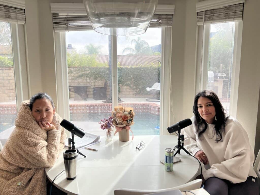 The Anatomy of Friendship. 1 Joanne and Nabiha at a round kitchen table with microphones, recording a podcast. In the cozy room, large windows reveal a pool in the backyard. A vase of flowers and some beverages are on the table.