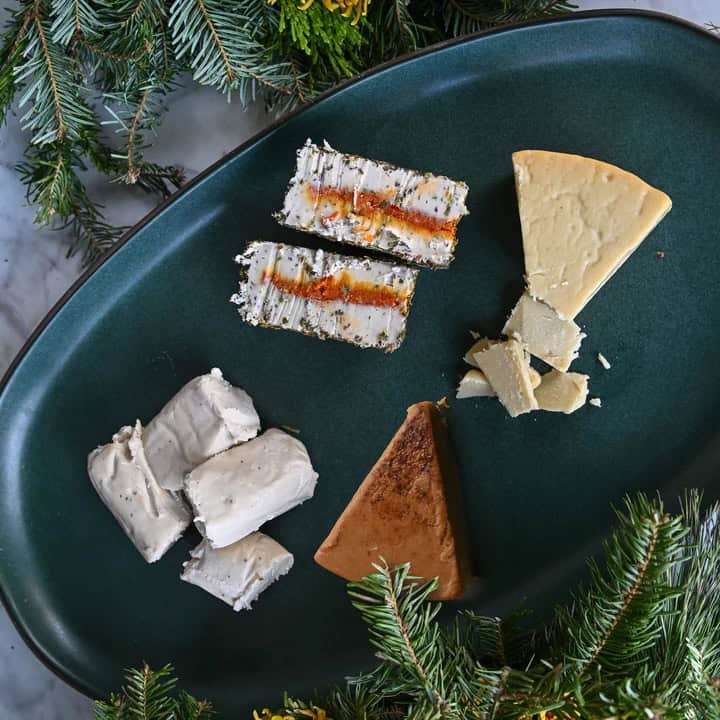 The Korean Vegan's 2025 VEGAN Holiday Gift GUIDE. 46 A dark green platter featuring assorted cheese selections, including wedges and slices with visible herbs and spices. Perfect for 2024, the platter is surrounded by green pine branches, enhancing its festive presentation—ideal for a holiday gift guide.