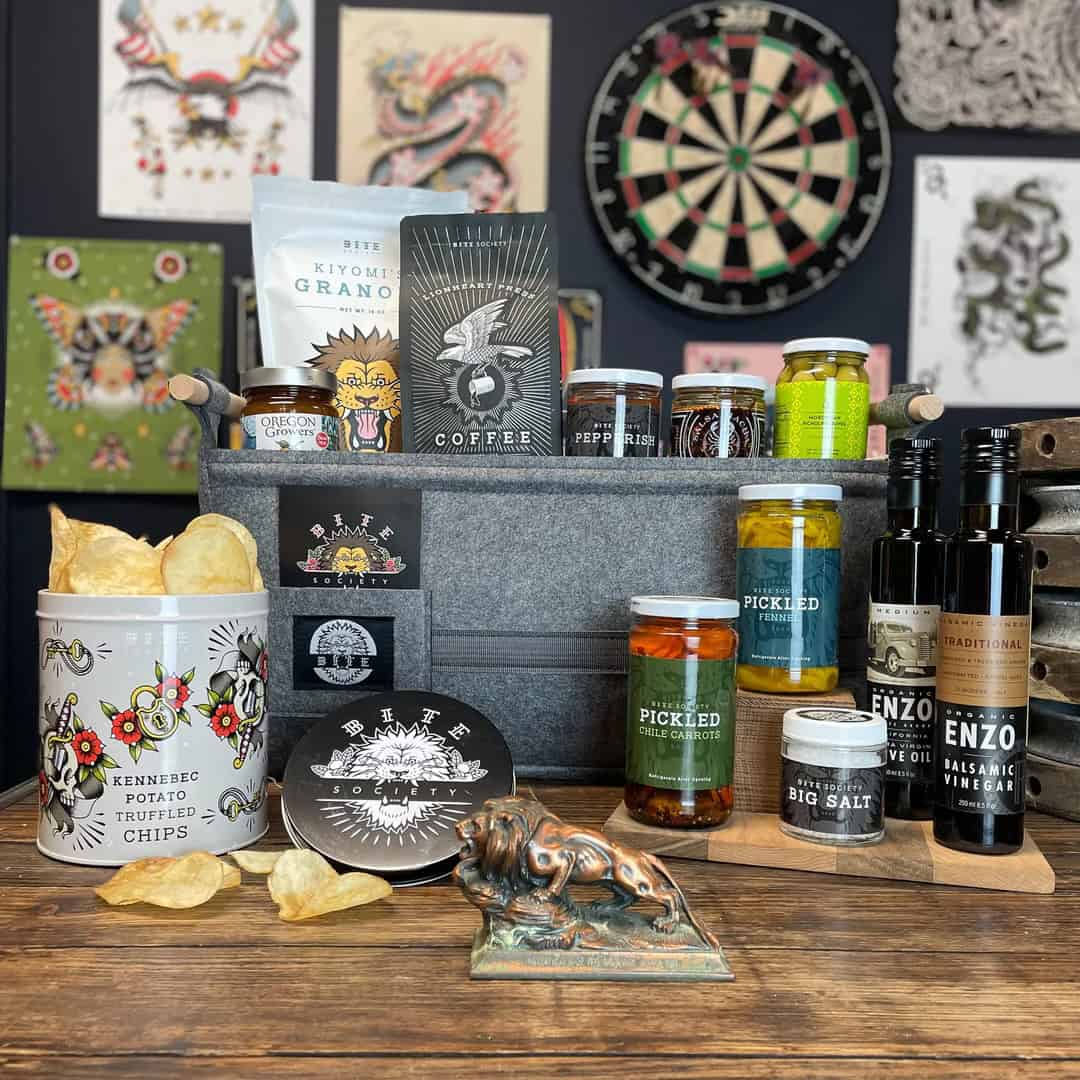 The Korean Vegan's 2025 VEGAN Holiday Gift GUIDE. 47 A gift basket on a wooden table features truffled potato chips, granola, coffee, pickled vegetables, salts, vinegars, and more—a perfect addition to any Holiday Gift Guide. Artistic wall hangings and a dartboard in the background add charm to this delightful scene.