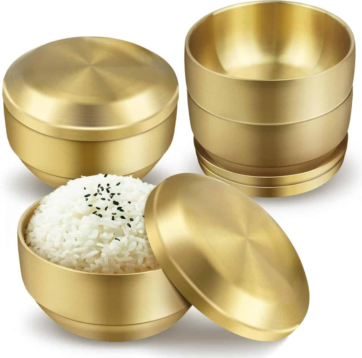 The Korean Vegan's 2025 VEGAN Holiday Gift GUIDE. 20 Shown are three gold-toned stainless steel bowls with lids, perfect as a stylish holiday gift. One bowl is filled with white rice garnished with black sesame seeds, while the others are stacked empty, showcasing their modern, sleek design.