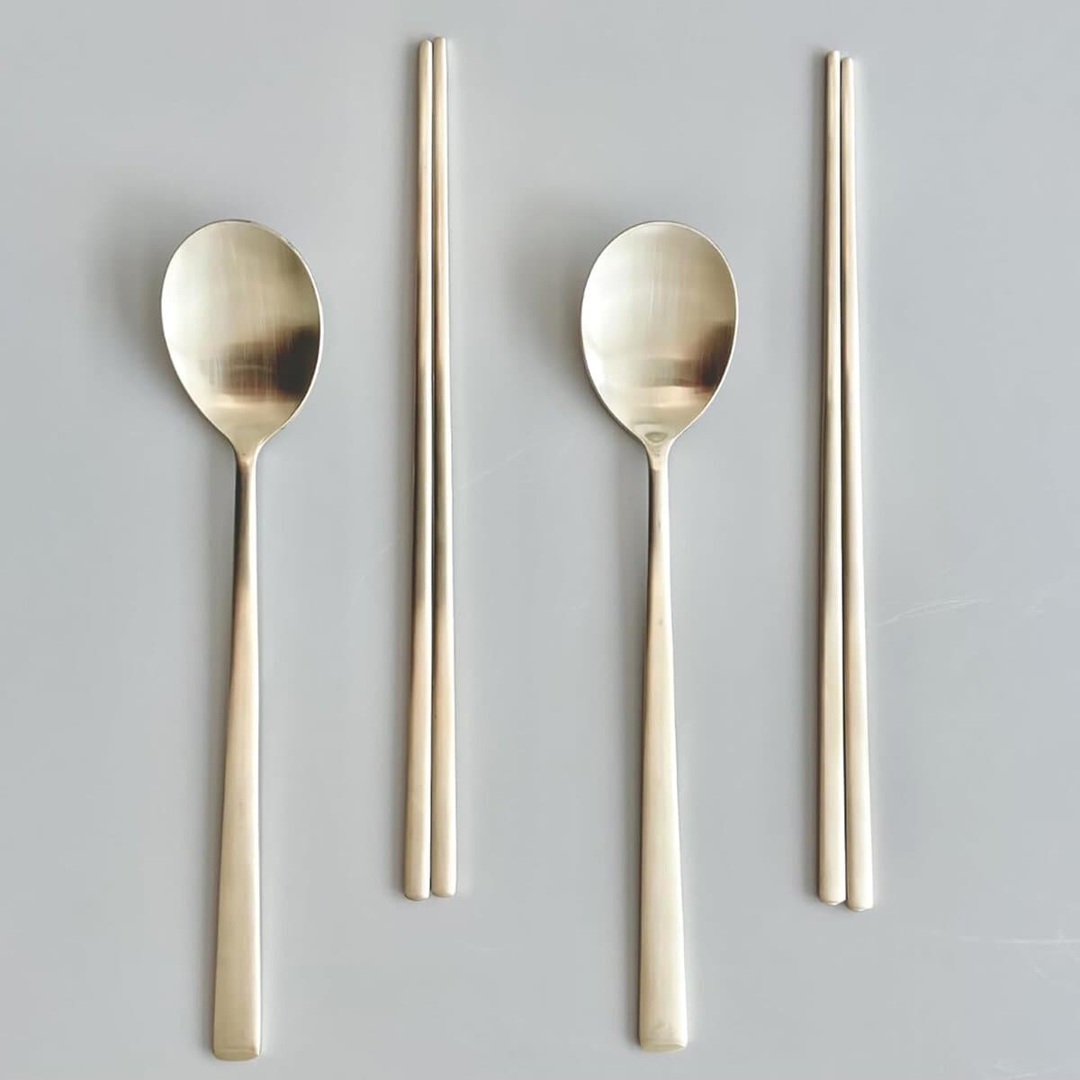 The Korean Vegan's 2025 VEGAN Holiday Gift GUIDE. 21 A neatly arranged set of two metal spoons paired with two pairs of metal chopsticks on a light grey background. The utensils have a sleek, modern design with a shiny finish.