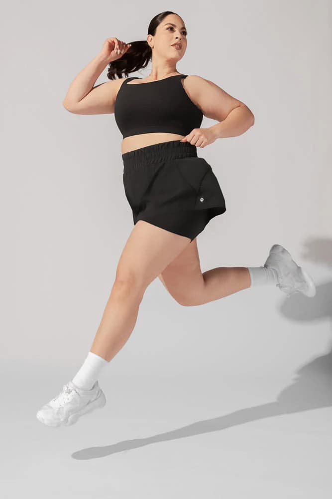 The Korean Vegan's 2025 VEGAN Holiday Gift GUIDE. 35 A person in a black sports bra and shorts jumps with energy and enthusiasm. They wear white sneakers and socks against a plain background, one arm bent and the other extended back.