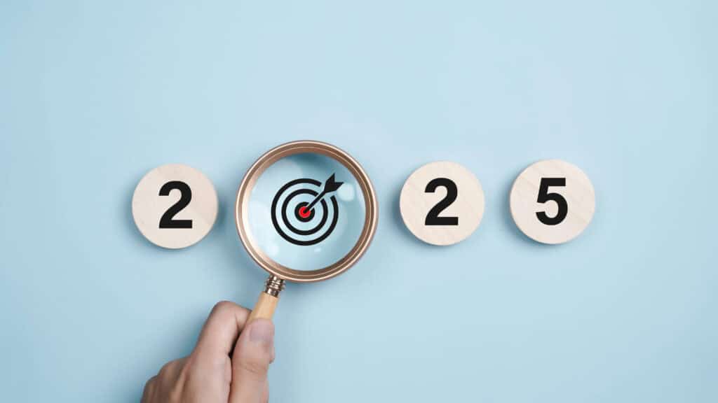 A Note on New Year's Resolutions 1 A hand with a magnifying glass zeroes in on a red and black target symbol nestled between the numbers 2025, embodying a New Years Resolution. Set against a light blue backdrop, the numbers rest on wooden circles, emphasizing focus and determination for the year ahead.