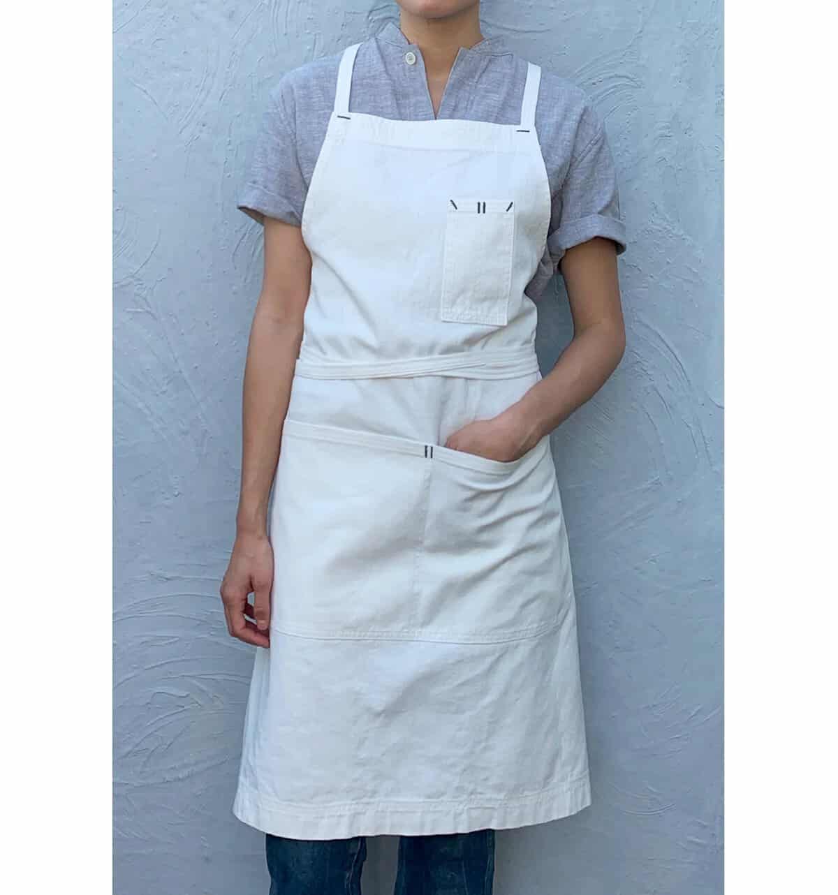 The Korean Vegan's 2025 VEGAN Holiday Gift GUIDE. 13 Standing against a textured gray wall, the person wears a short-sleeved gray shirt and a white apron with pockets. One hand rests thoughtfully in the apron pocket.