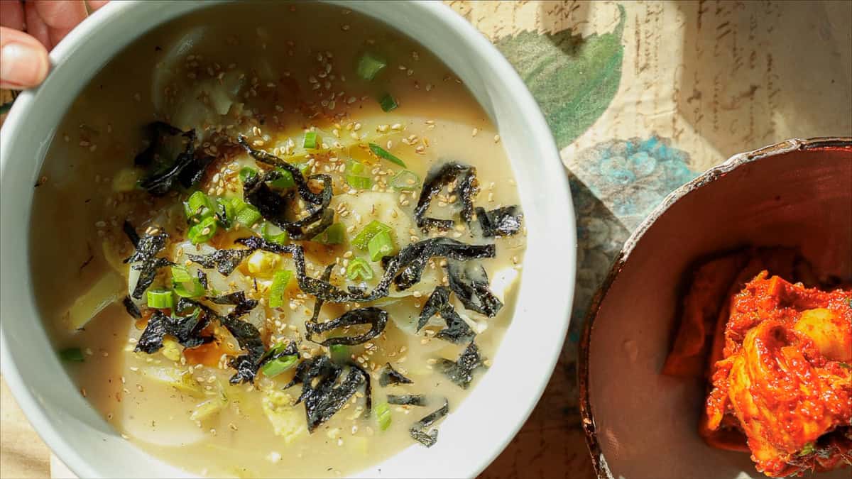 Delicious Vegan Rice Cake Soup (Tteokguk) with Dumplings. 1 A bowl of steaming vegan rice cake soup with sliced vegetables, sprinkled with seaweed, sesame seeds, and green onions, sits on a textured surface. Beside it is a dish of vibrant red kimchi, partially visible.