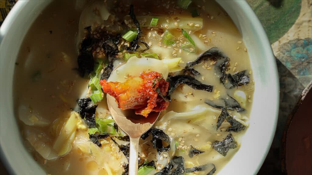 Delicious Vegan Rice Cake Soup (Tteokguk) with Dumplings. 4 A bowl of savory vegan rice cake soup with sliced seaweed, chopped green onions, sesame seeds, and a wooden spoon holding a dollop of spicy sauce. The soup has a creamy broth with thin noodles, creating a hearty and flavorful dish.
