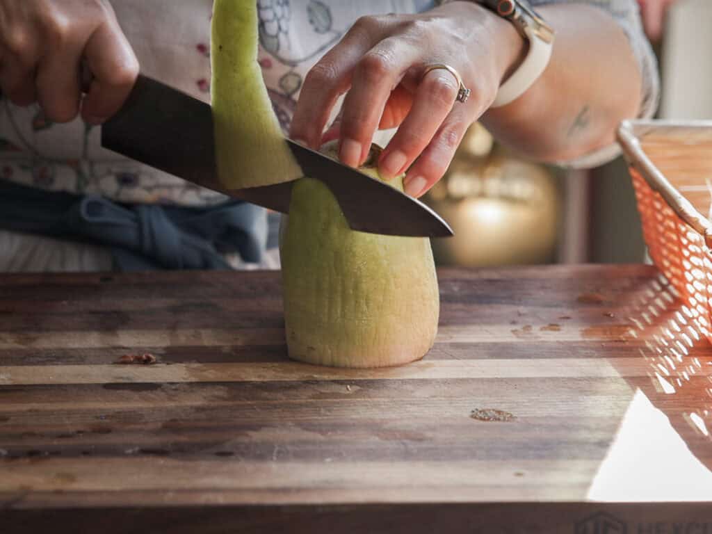 Delicious Vegan Rice Cake Soup (Tteokguk) with Dumplings. 14 A person slicing the peel of a Korean radish on a wooden cutting board. They are using a large knife and wearing a smartwatch and a ring. The vegetable is upright, partially peeled, with a basket in the background.