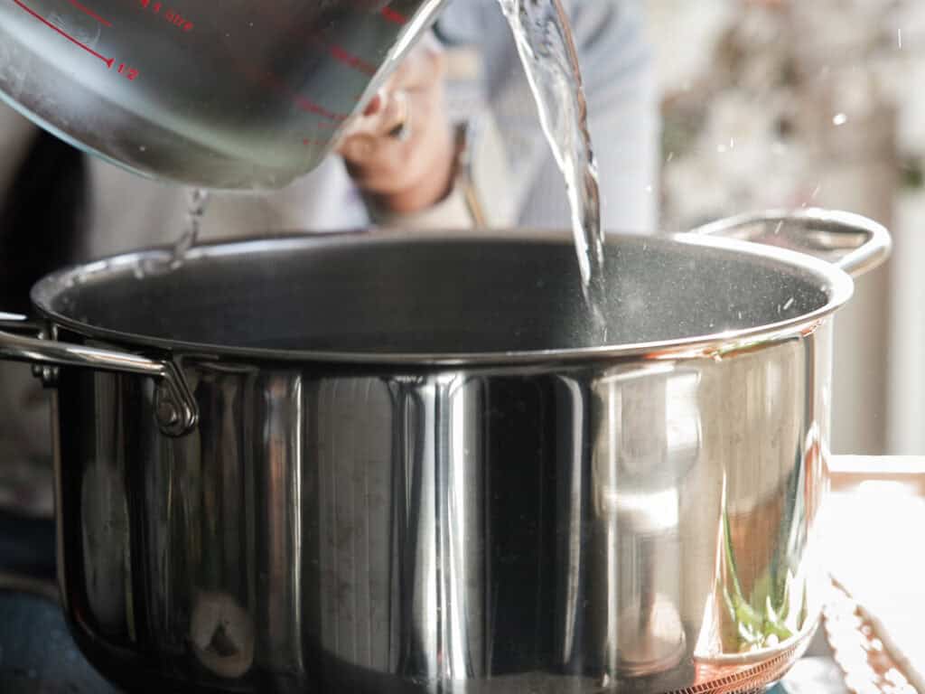 Delicious Vegan Rice Cake Soup (Tteokguk) with Dumplings. 18 A person is pouring water from a large measuring cup into a shiny stainless steel pot on a stove. The setting appears to be a kitchen, with blurred background details.