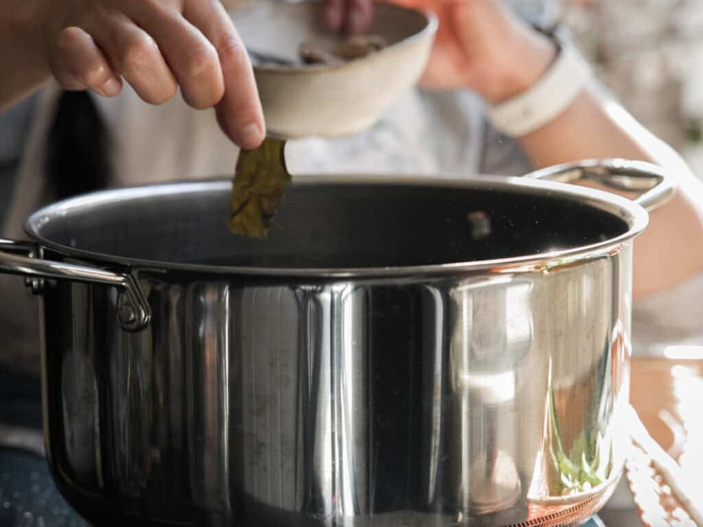 Delicious Vegan Rice Cake Soup (Tteokguk) with Dumplings. 19 A person adds ingredients from a small bowl into a large stainless steel pot. The focus is on the hand holding a leaf above the pot. The scene suggests cooking or preparing a dish.
