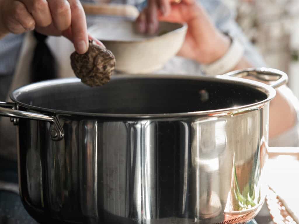 Delicious Vegan Rice Cake Soup (Tteokguk) with Dumplings. 20 A person holds a mushroom and a bowl over a shiny metal pot on a stove, suggesting cooking or preparing a meal. The focus is on the mushroom and pot, with soft natural light illuminating the scene.
