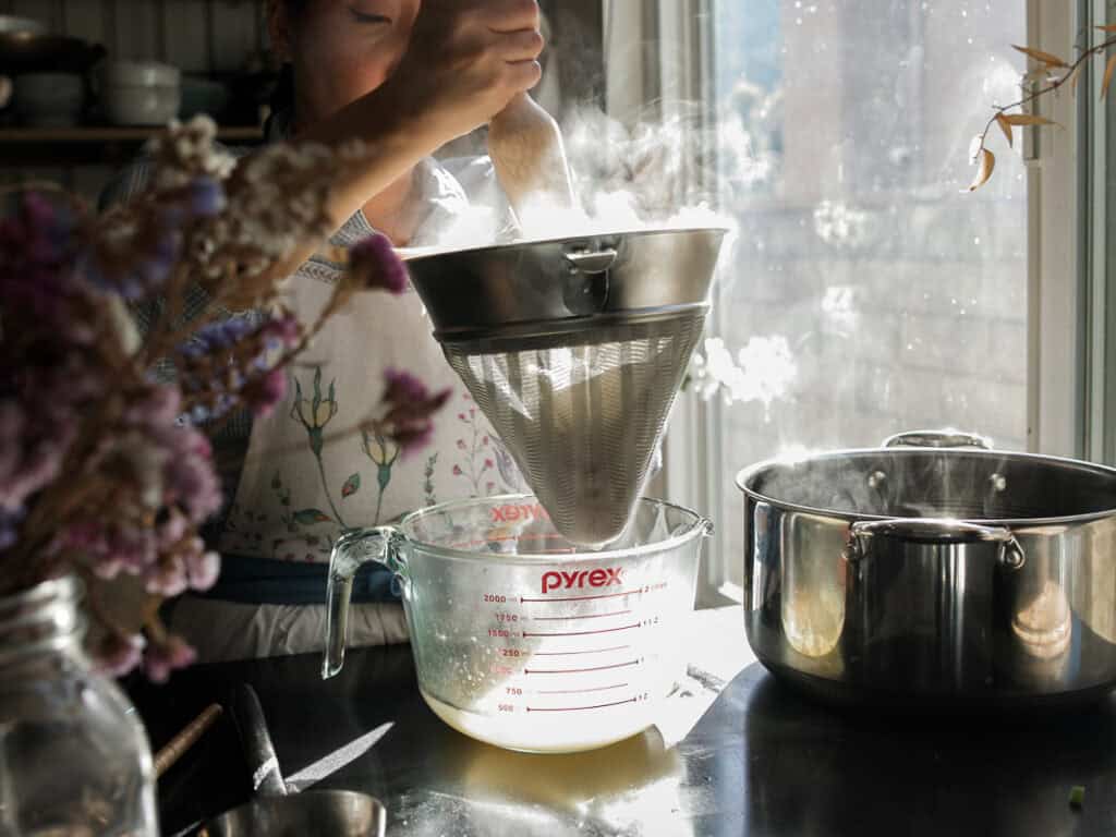 Delicious Vegan Rice Cake Soup (Tteokguk) with Dumplings. 23 A person strains hot broth through a metal sieve into a glass measuring cup. Sunlight streams through a nearby window, illuminating steam rising from the pot. A bouquet of flowers sits in the foreground.