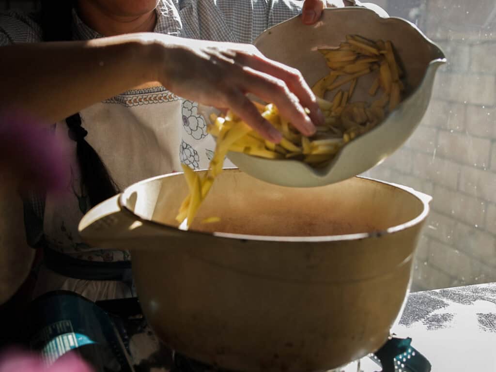 Delicious Vegan Rice Cake Soup (Tteokguk) with Dumplings. 29 A person wearing an apron is adding sliced potatoes from a bowl into a large pot. Sunlight is streaming through a window, casting a warm glow over the scene.