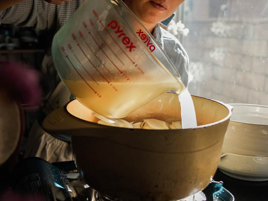 Delicious Vegan Rice Cake Soup (Tteokguk) with Dumplings. 30 A person pouring liquid from a Pyrex measuring cup into a pot on a stove. The bright natural light from a window illuminates the scene, highlighting the kitchen setting.