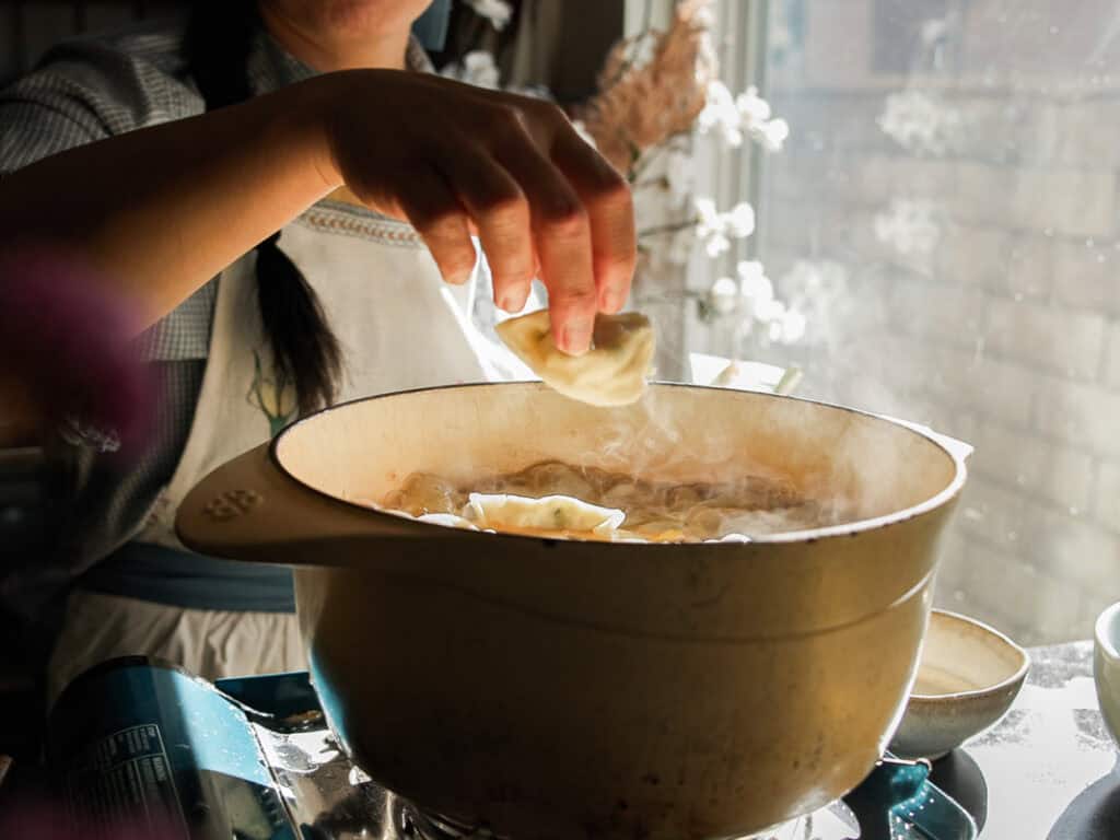 Delicious Vegan Rice Cake Soup (Tteokguk) with Dumplings. 33 A person is cooking dumplings in a steaming pot on a stovetop. One hand is holding a dumpling above the pot. Sunlight streams through a nearby window, illuminating the kitchen area.