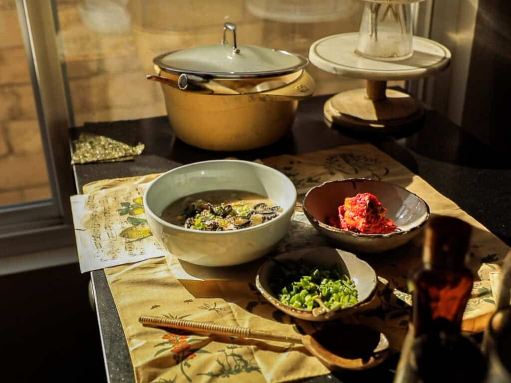 Delicious Vegan Rice Cake Soup (Tteokguk) with Dumplings. 36 A sunlit table with a bowl of soup and a red dish, surrounded by a small bowl of chopped green onions, a metallic pot with a lid, and a wooden utensil. A yellow printed cloth covers the table surface.