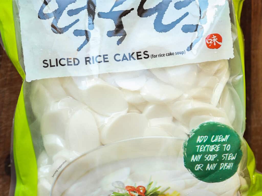 Delicious Vegan Rice Cake Soup (Tteokguk) with Dumplings. 6 A package of sliced rice cakes for soup, with clear plastic showing white oval slices inside. The green and red label suggests adding them to soups or stews for a chewy texture. The top of the package has stylized text.