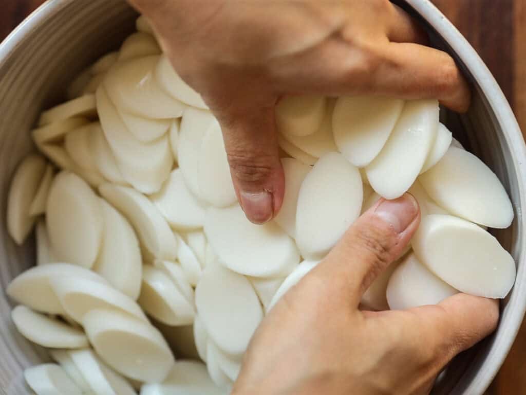 Delicious Vegan Rice Cake Soup (Tteokguk) with Dumplings. 26 Hands holding oval-shaped, sliced rice cakes in a bowl, likely being prepared for cooking. The rice cakes are smooth and white, filling the bowl completely. The background is a wooden surface.