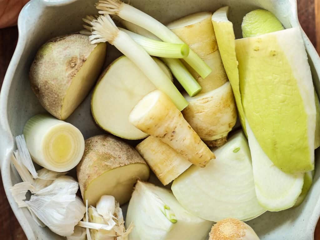 Delicious Vegan Rice Cake Soup (Tteokguk) with Dumplings. 11 A bowl filled with various chopped vegetables, including potatoes, leeks, carrot, radish peel, and garlic cloves, all arranged closely together.