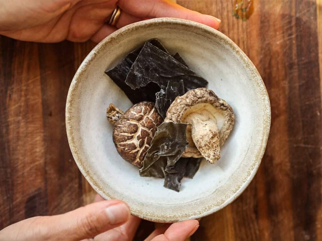 Delicious Vegan Rice Cake Soup (Tteokguk) with Dumplings. 10 Hands holding a ceramic bowl containing dried shiitake mushrooms and seaweed, set on a wooden surface.