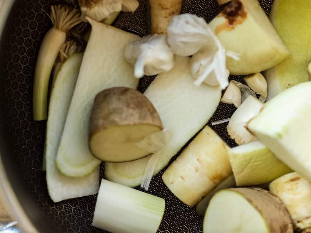 Delicious Vegan Rice Cake Soup (Tteokguk) with Dumplings. 16 A close-up of various chopped vegetables in a pan, including potatoes, onion, garlic, leek. The ingredients are in preparation for cooking, with visible skin and varied textures in a black pan.