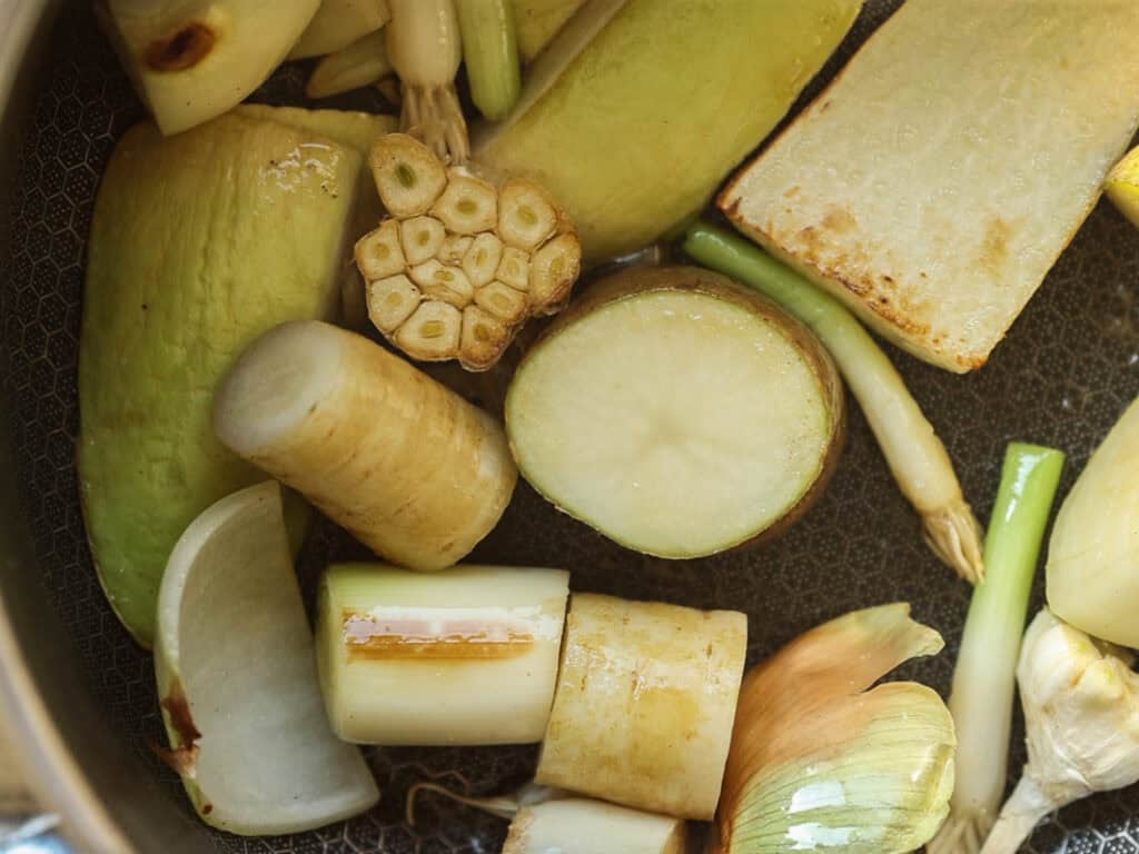 Delicious Vegan Rice Cake Soup (Tteokguk) with Dumplings. 17 Chopped vegetables, including garlic, leeks, onion, and other root vegetables, are scattered in a pan. They are partially sautéed and vary in size and color, with textures ranging from smooth to slightly charred.