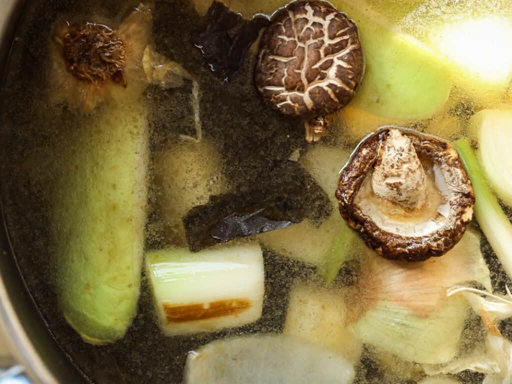 Delicious Vegan Rice Cake Soup (Tteokguk) with Dumplings. 13 A pot filled with a variety of vegetables and mushrooms, including shiitake mushrooms, leeks, and seaweed, submerged in a clear broth. The ingredients are partially cooked, visible beneath the surface of the liquid.
