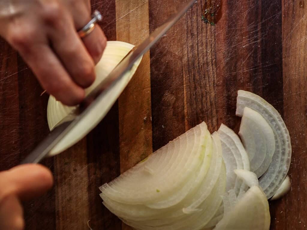 Delicious Vegan Rice Cake Soup (Tteokguk) with Dumplings. 8 A person slices an onion on a wooden cutting board. The left hand holds the onion steady, while the right hand uses a knife to cut even slices. The onion pieces are arranged on the cutting board.