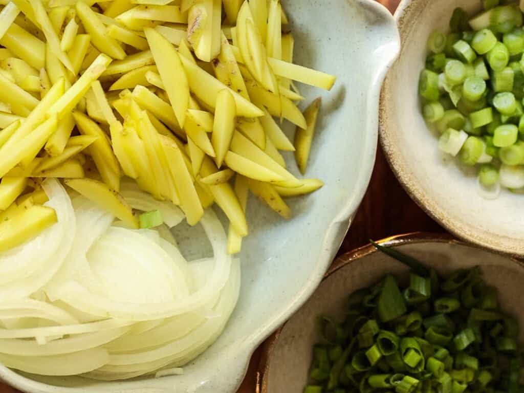 Delicious Vegan Rice Cake Soup (Tteokguk) with Dumplings. 5 Close-up of a bowl containing julienned potato, another with sliced onions, and a third with chopped green onions. The ingredients are fresh and vibrant, displayed in ceramic bowls on a wooden surface.