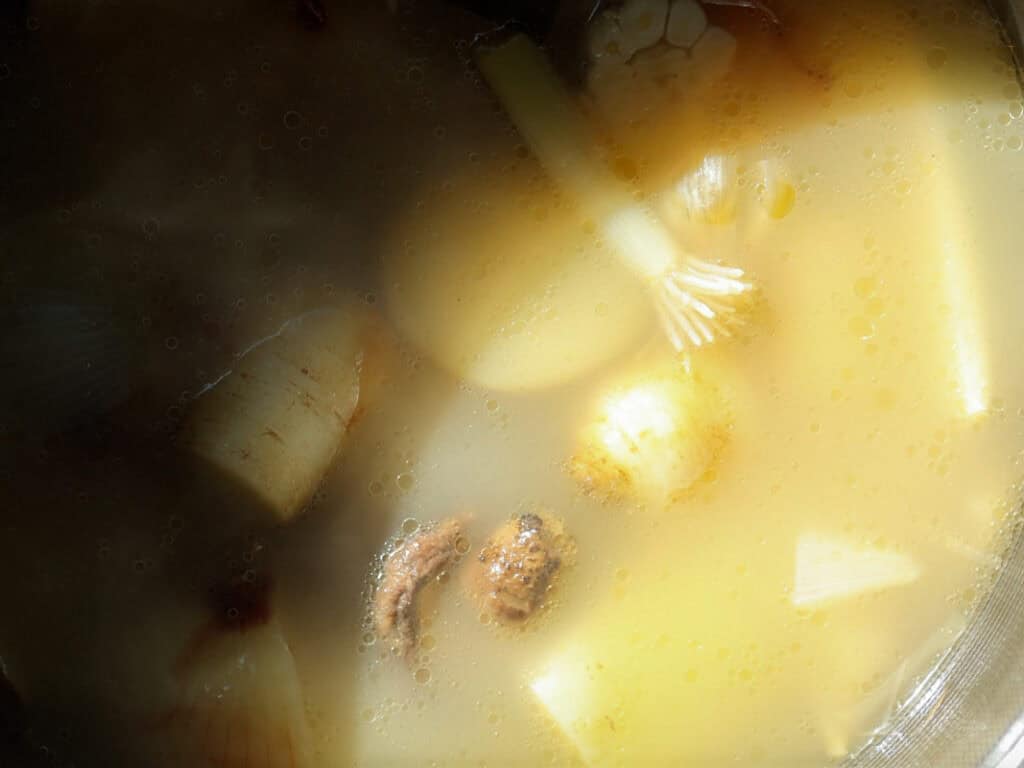 Delicious Vegan Rice Cake Soup (Tteokguk) with Dumplings. 22 Close-up of a simmering soup with visible ingredients, including potatoes, onions, scallions, and other vegetables, in a rich, light-colored broth, partially lit by sunlight.