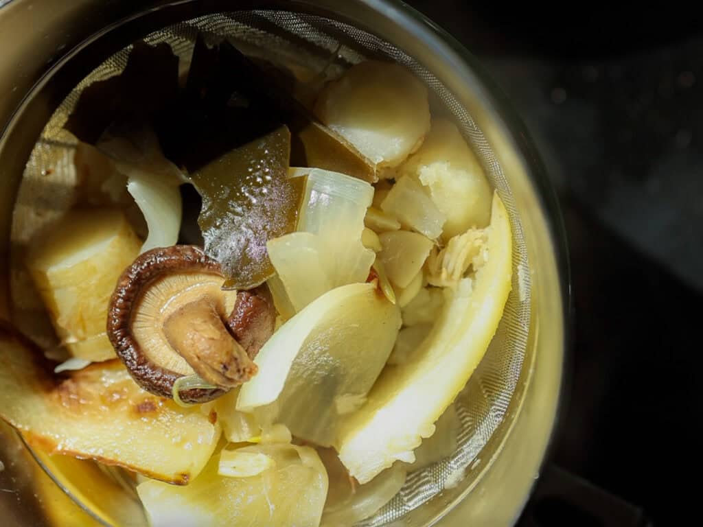 Delicious Vegan Rice Cake Soup (Tteokguk) with Dumplings. 24 A close-up of a metal strainer containing various cooked vegetables, including sliced carrot, onions, and a whole mushroom, with visible steam and a warm light illuminating the ingredients.