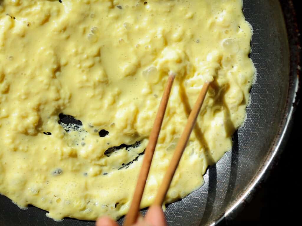 Delicious Vegan Rice Cake Soup (Tteokguk) with Dumplings. 12 Close-up of scrambled vegan eggs being stirred with chopsticks in a non-stick frying pan. The eggs are partially cooked, with a creamy texture and pale yellow color, indicating they are still in the process of being prepared.