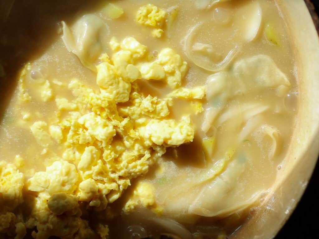 Delicious Vegan Rice Cake Soup (Tteokguk) with Dumplings. 35 A close-up of a bowl of creamy vegan rice cake soup containing scrambled vegan eggs, dumplings, and sliced onions. The broth appears rich and thick, with a golden hue and pieces of cooked egg floating on the surface.