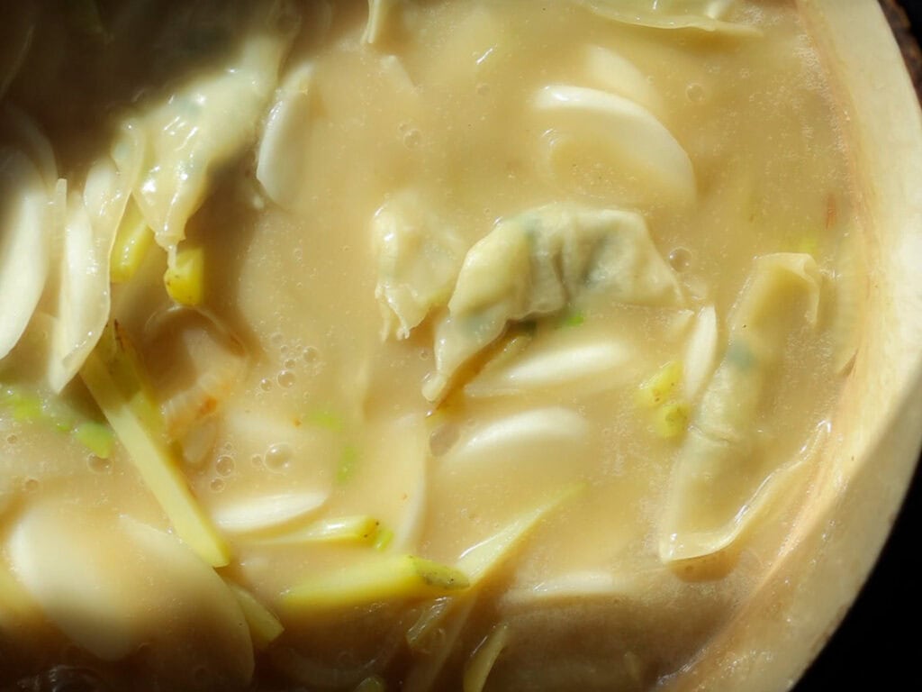 Delicious Vegan Rice Cake Soup (Tteokguk) with Dumplings. 34 A close-up of a creamy vegan rice cake soup with dumplings and sliced vegetables. The mixture appears thick, with visible pieces of dumplings and green onion slices, all enveloped in a smooth, light-colored broth.