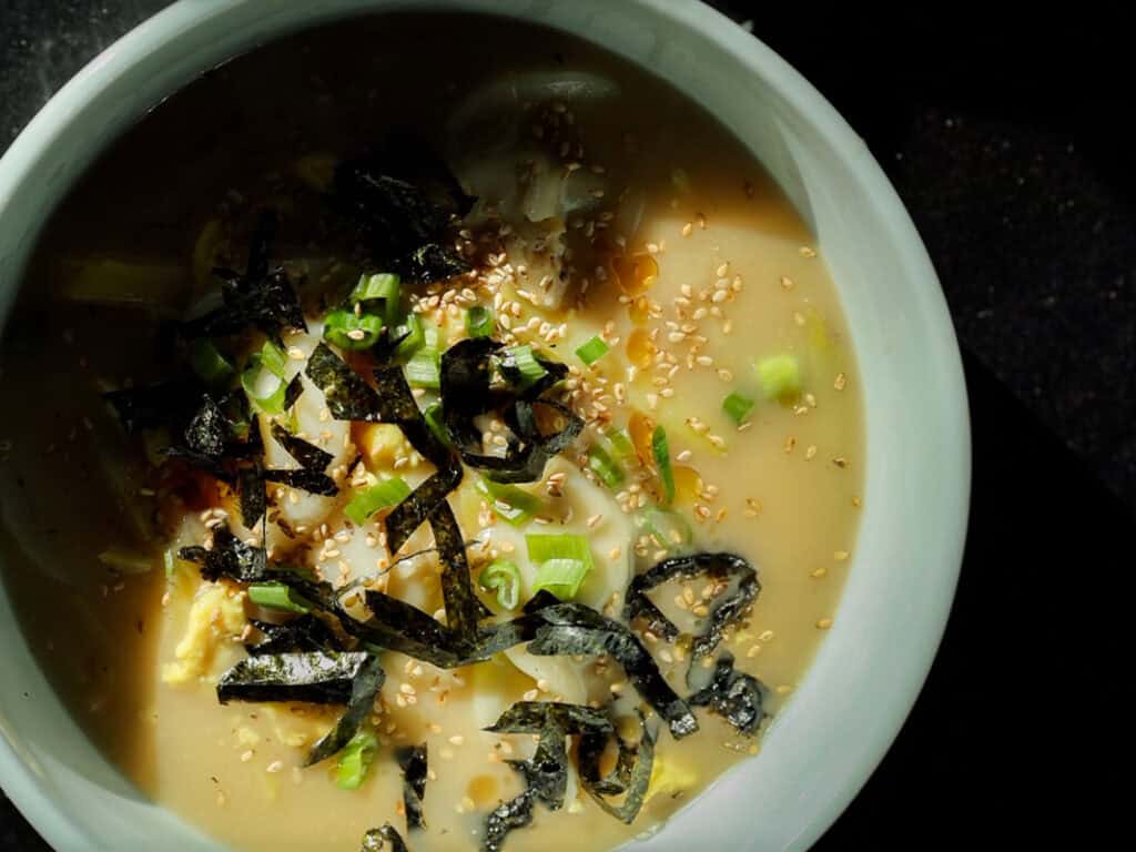 Delicious Vegan Rice Cake Soup (Tteokguk) with Dumplings. 2 A close-up of a bowl of vegan rice cake soup in a creamy broth, topped with sliced green onions, seaweed strips, sesame seeds, and what appears to be an egg. The bowl is set against a dark background with natural lighting casting shadows.