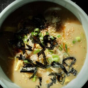 Delicious Vegan Rice Cake Soup (Tteokguk) with Dumplings. 37 A bowl of creamy vegan rice cake soup topped with seaweed strips, chopped green onions, and sesame seeds, with dumplings. The dish is set against a dark background.