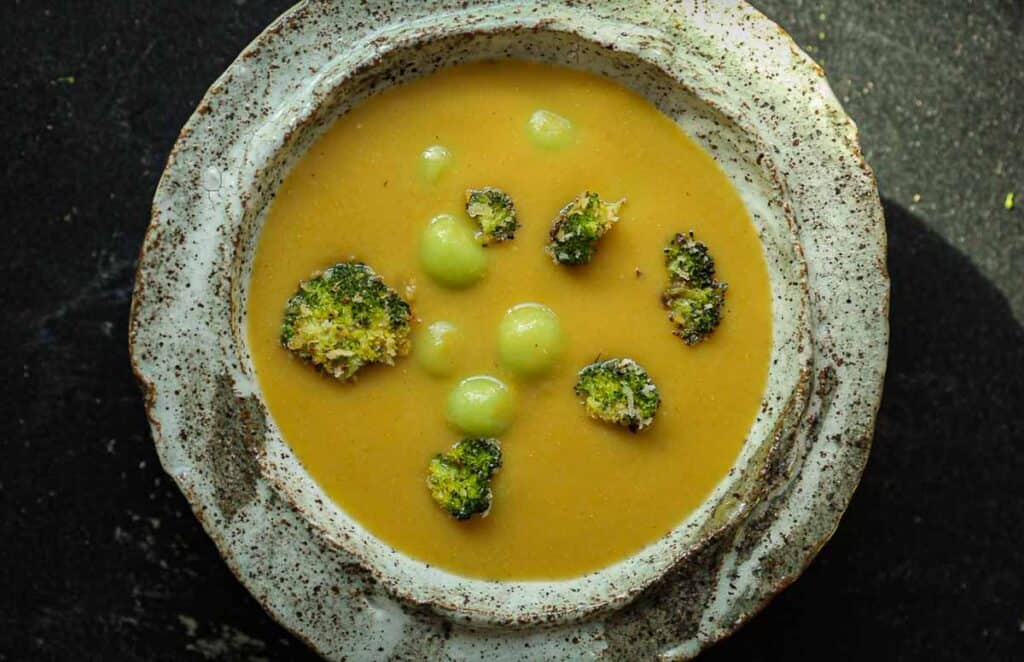Creamy Broccoli Soup with Dumplings - delicious and Gluten-Free! 2 A speckled ceramic bowl filled with creamy broccoli soup garnished with broccoli florets and small green spheres. The bowl is set against a dark background.