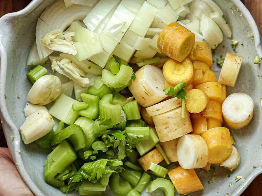 Creamy Broccoli Soup with Dumplings - delicious and Gluten-Free! 4 A bowl filled with chopped vegetables, including onions, yellow carrots, garlic cloves, and celery, arranged in sections. The ingredients are fresh and colorful, highlighting a mix of greens, yellows, and whites.
